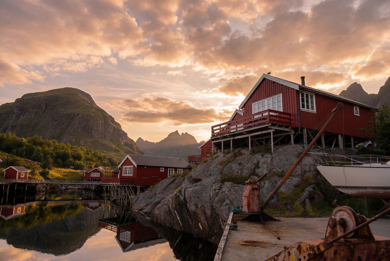 Rote Fischerhäuser von Sørvågen reihen sich am Fjordufer, das ruhige Wasser spiegelt die bunten Holzbauten, im Hintergrund norwegische Natur.