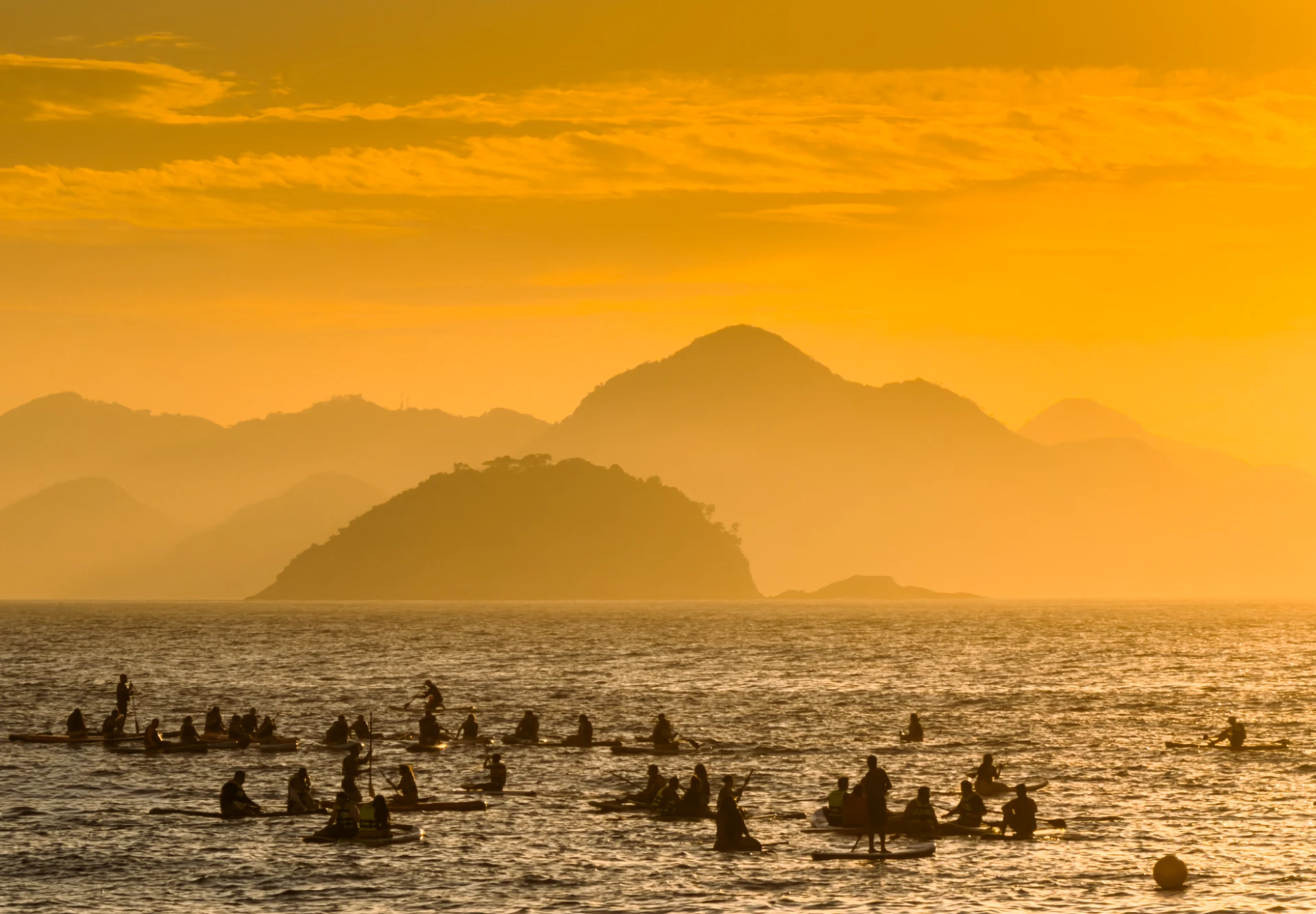 Gruppe von Paddlern auf dem Meer bei Sonnenaufgang mit warmem Goldton, Silhouetten vor Bergen und Inseln in Rio de Janeiro.