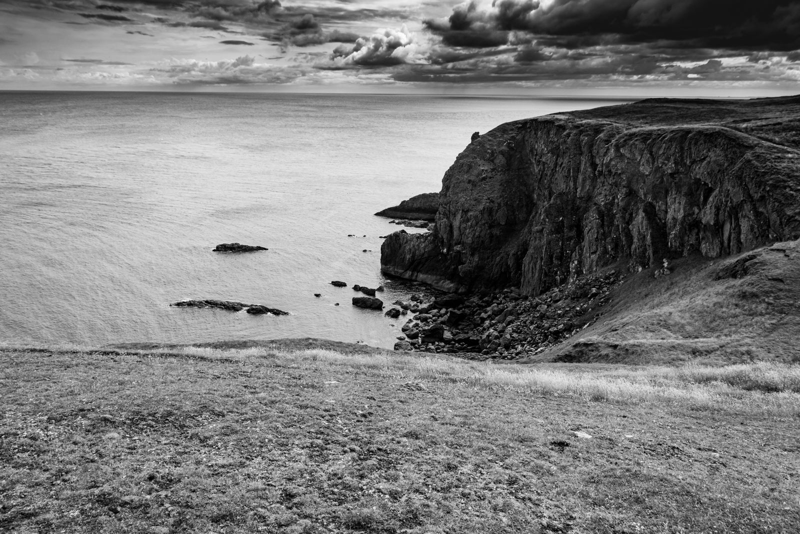Schwarzweiß-Fotografie von dramatischen Felsklippen der Isle of Skye, ruhige See im Vordergrund, weite Wolken am Himmel.