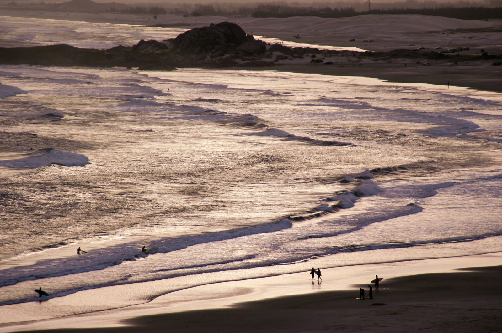 Ein Surfer mit Board läuft am brasilianischen Strand entlang. Sanfte Wellen, untergehende Sonne und vielfältige Farben prägen die ruhige Szene.