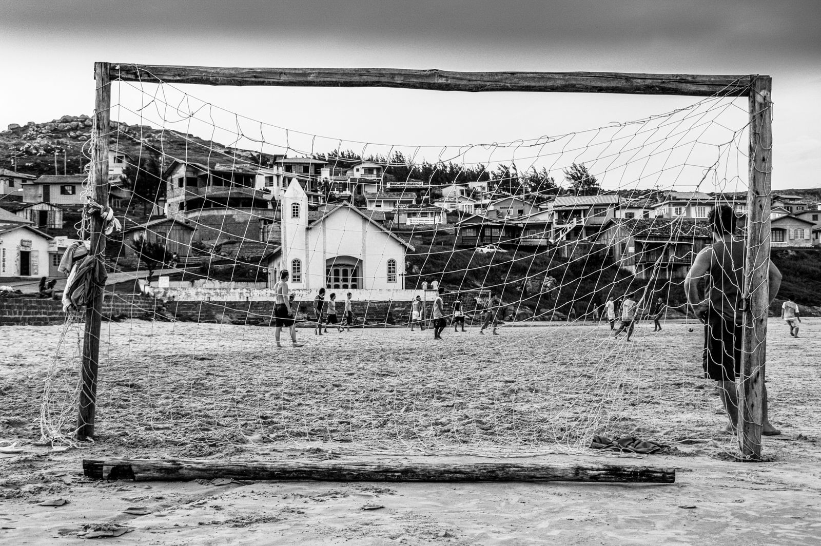 Kinder spielen barfuß Fußball am improvisierten Sandtor aus Holzpfosten an einem kleinen brasilianischen Strand bei warmem Sonnenlicht.
