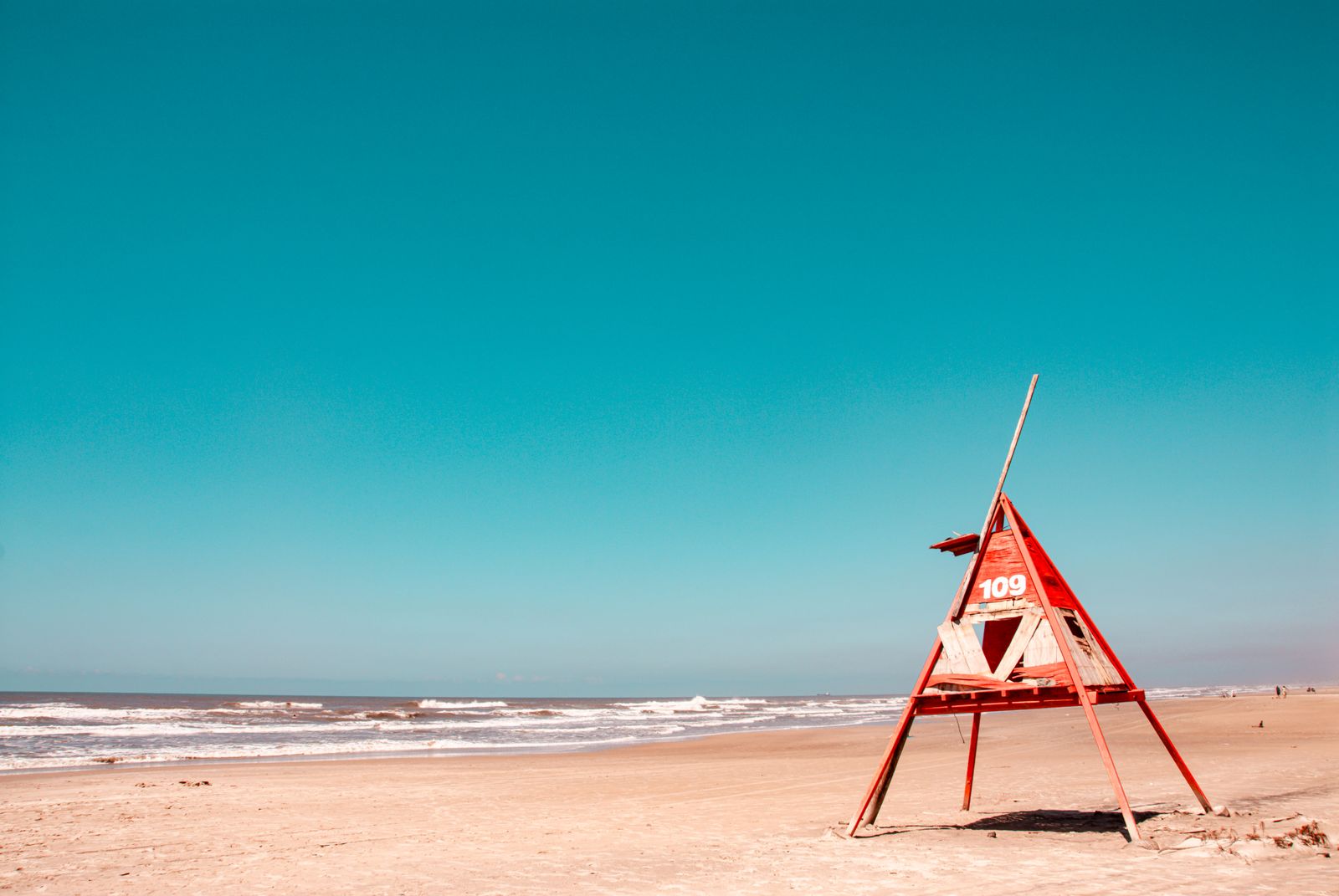 Ein moderner Rettungsturm steht am ruhigen, brasilianischen Sandstrand unter klarem Himmel. Minimalistische, vielfältige Farben.