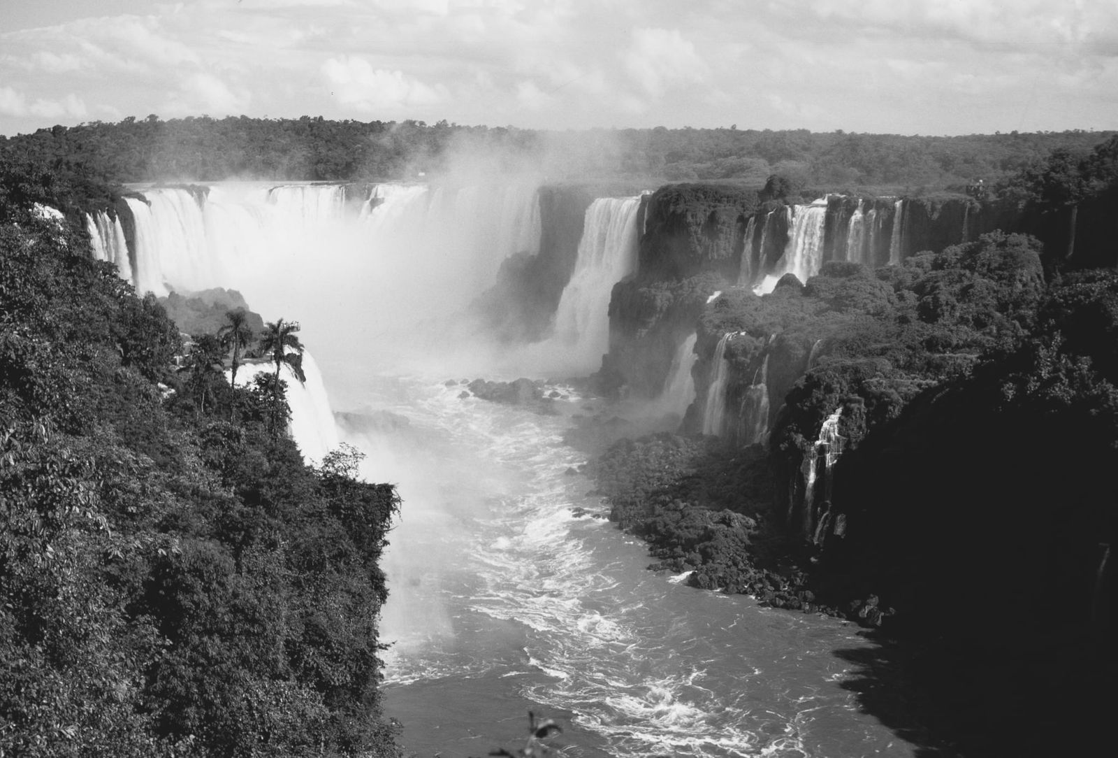 Blick auf die stufenförmigen Iguazú Wasserfälle, sanftes Licht fällt durch dichtes Blattwerk, feiner Sprühnebel über den Wassermassen.