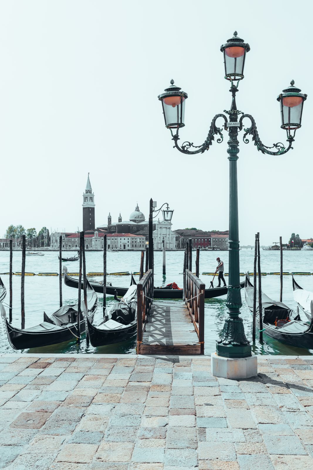 Mehrere Gondeln schaukeln auf ruhigem Wasser bei Venedig. Im Vordergrund steht eine historische Laterne, die das Bild sanft beleuchtet.