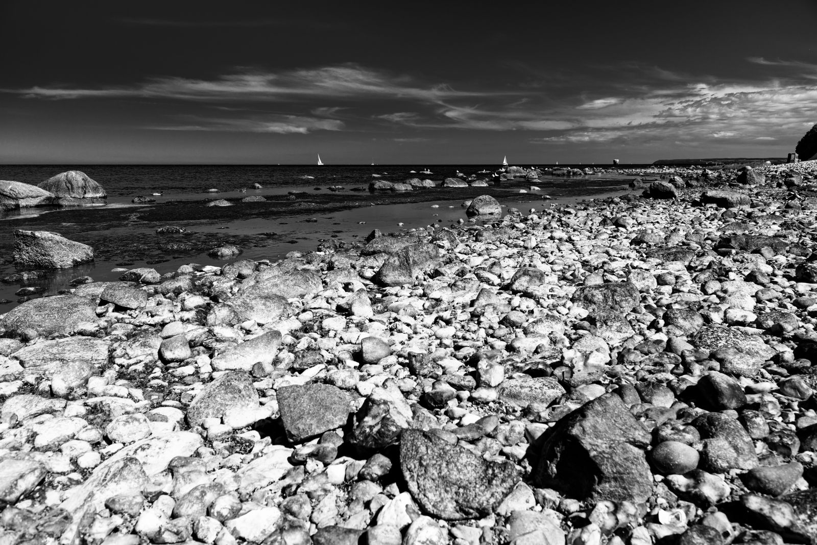 Große Felsbrocken liegen auf sandigem Strand an der Küste von Arkona, dahinter sanfte Wellen und ein weiter Himmel.