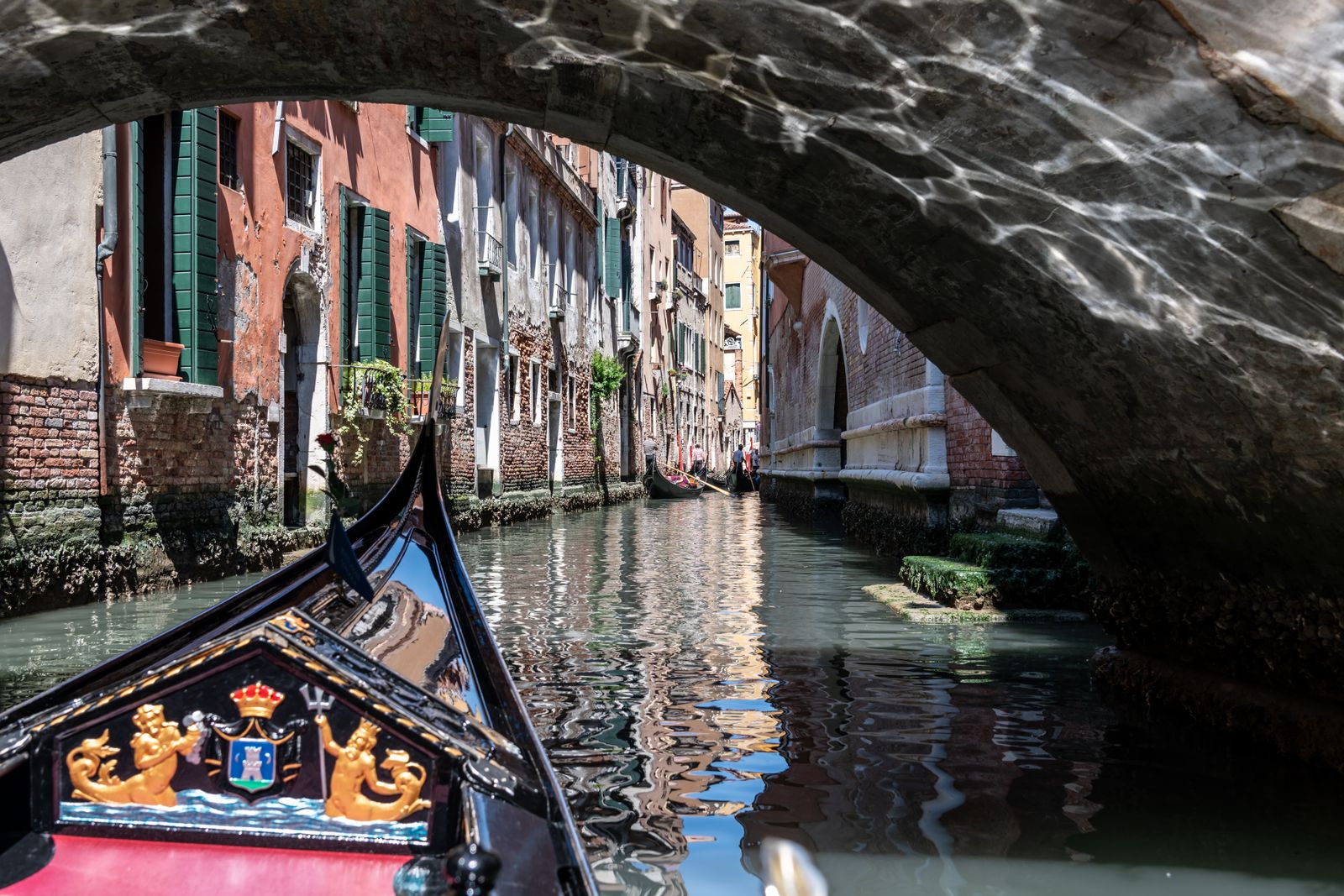 Eine Gondel fährt unter einer kunstvoll geschwungenen Brücke in San Marco hindurch, das Wasser spiegelt Lichtreflexe, urbanes Stadtmotiv.