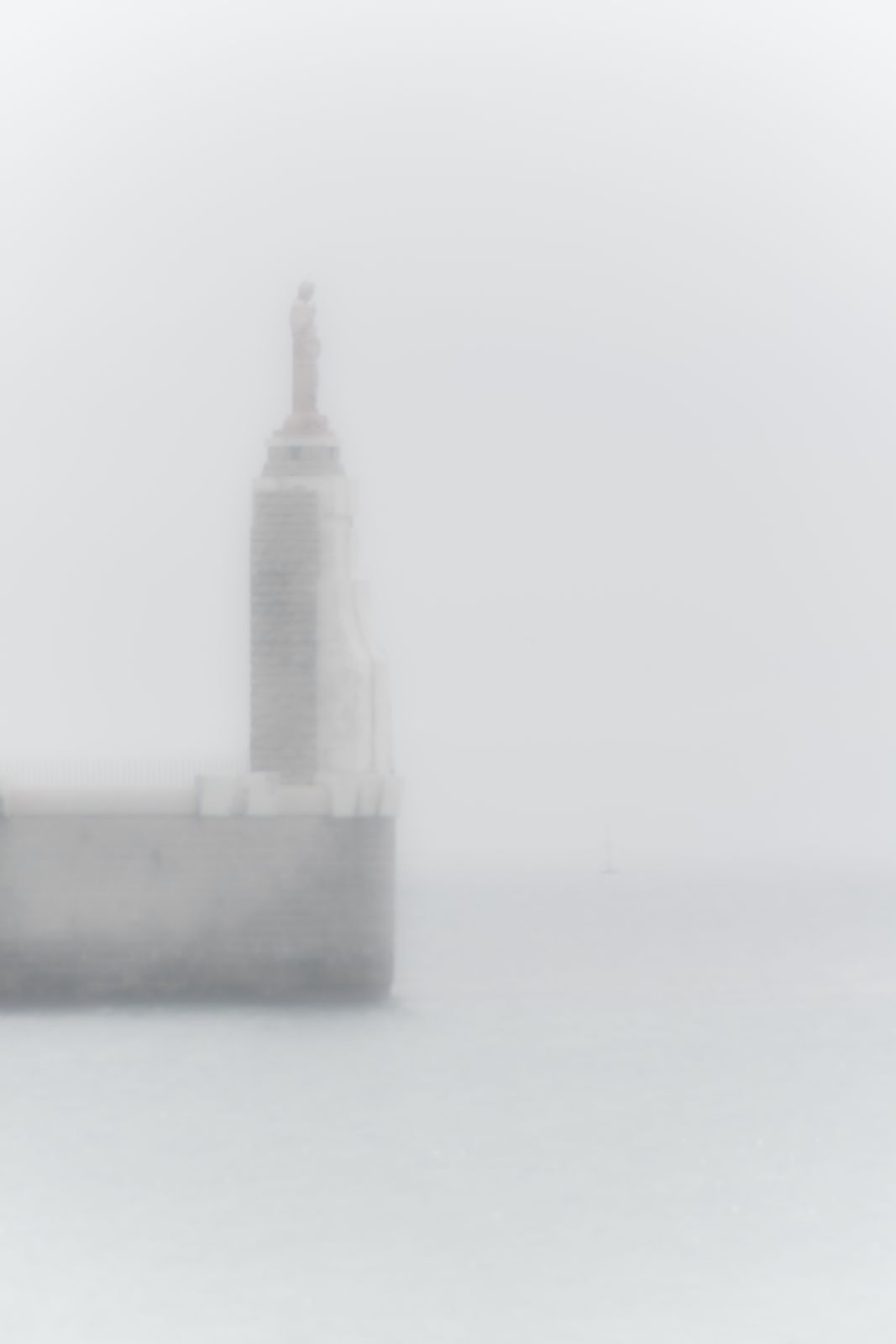 Ein Leuchtturm an der Hafeneinfahrt von Marseille ragt im Nebel empor, auf der Spitze eine Statue, minimalistisch und atmosphärisch inszeniert.