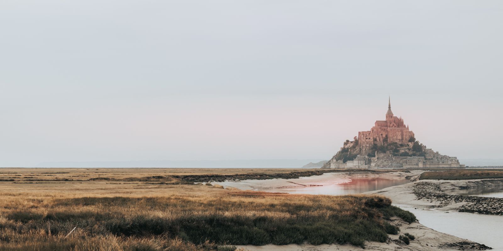 Das Panorama zeigt Mont-Saint-Michel im sanften, goldenen Licht, das Kloster erhebt sich ruhig über dem Watt, umgeben von vielfältigen Farben.