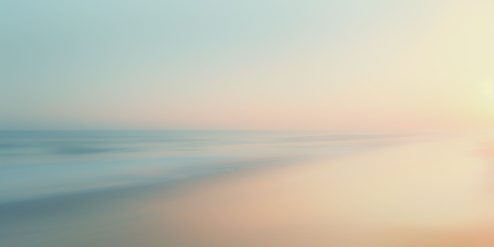 Zarter Sonnenaufgang über ruhigem Ozean mit feinem Sandstrand und weichem, farbenfrohem Himmel im Panorama-Format.