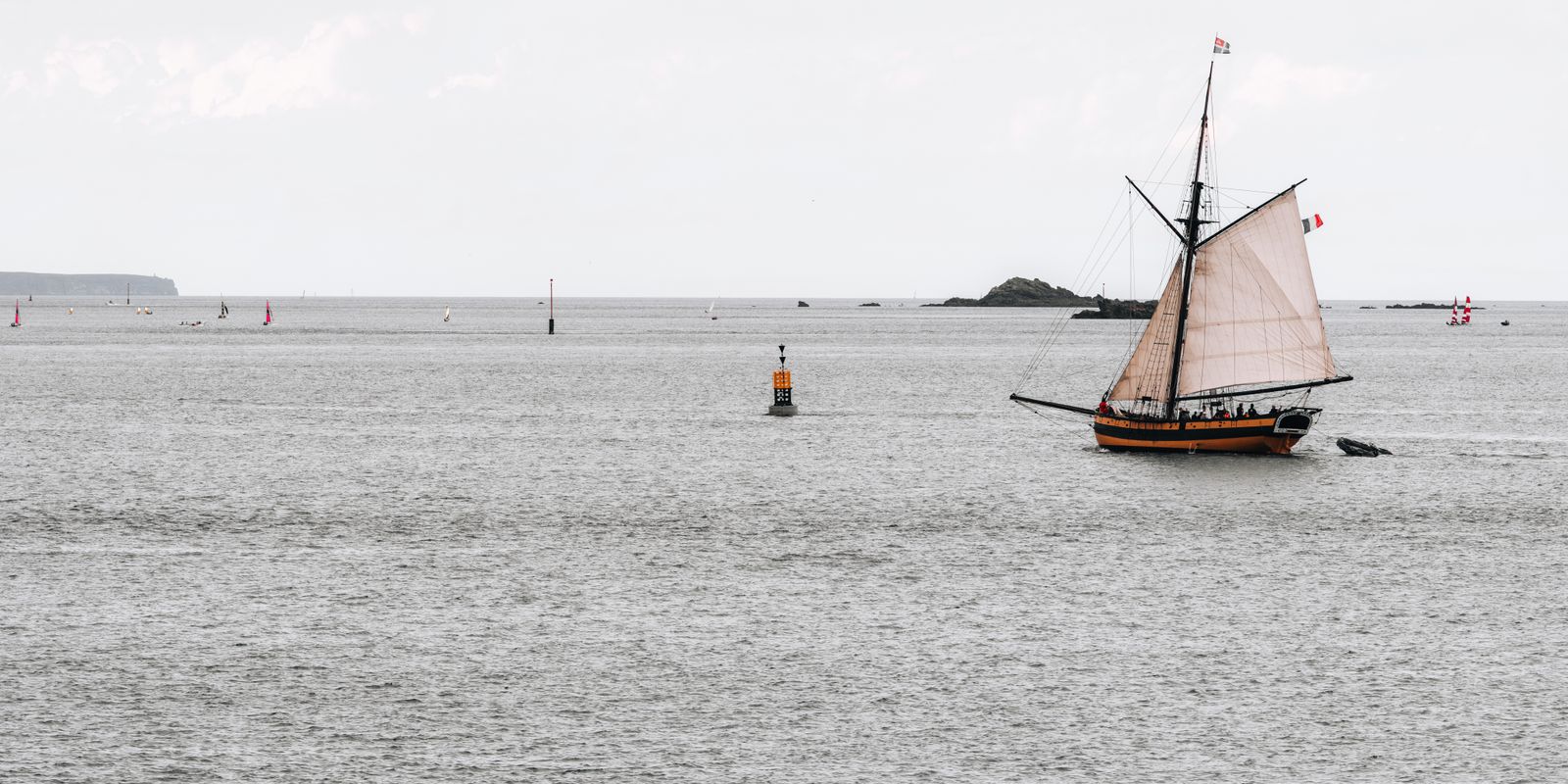 Historisches Segelschiff mit vollen Segeln fährt ruhig vor der felsigen Küste der Bretagne auf dem Meer, maritimes Motiv bei vielfältigem Licht.