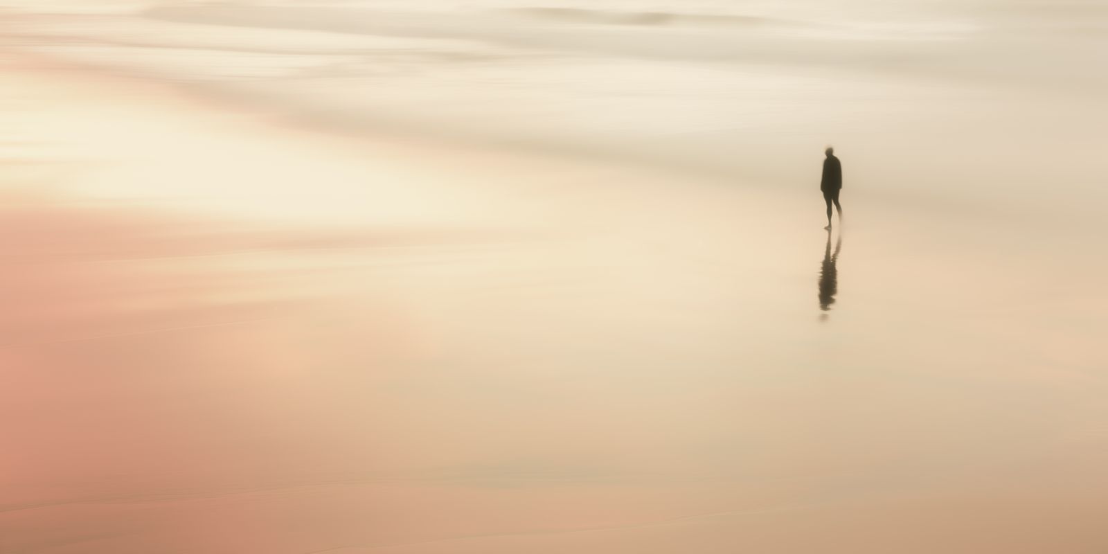 Eine einzelne menschliche Silhouette steht am ruhigen Strand, umgeben von warmen Sandtönen und einem farbenfrohen Himmel im Panoramaformat.